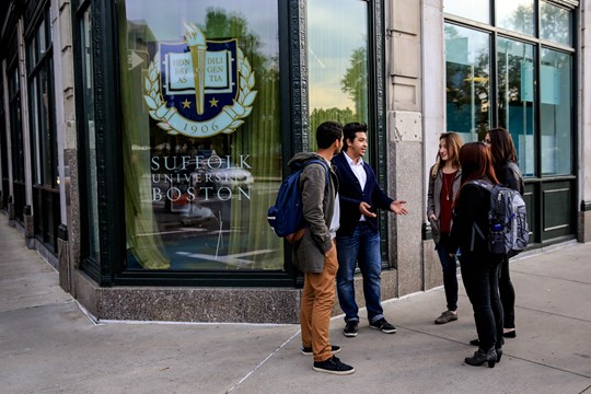 Five international students from INTO Suffolk University stand and converse in street corner in front of Suffolk building window in downtown Boston. 