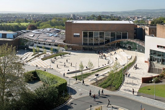 Aerial view of University of Exeter Forum, with wavy roof featuring triangular patterns and sloped quad with students on steps outside.