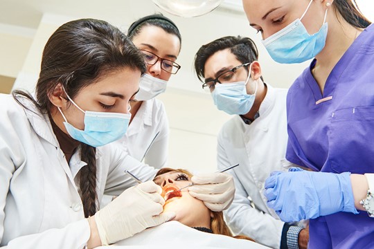 Group of dental students and instructor at Saint Louis University wear masks, white coats, and latex gloves as they work on patient’s teeth.