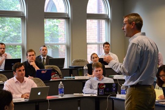 Saint Louis University students sit at rows of desks with laptops and listen to professor’s lecture in classroom.