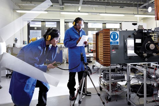 Two Manchester Metropolitan University students conduct experiment wearing sound-proof ear muffs, goggles, and blue coats in physics laboratory. 