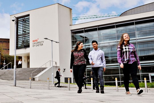 Three international students walk in front of INTO University of Exeter Centre building, with windows featuring INTO roundel.