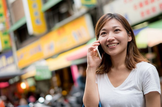 International student speaks on smartphone in front of colorful billboards in outdoor market.