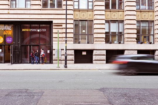 Street view of INTO Manchester Centre in Bridgewater House in the heart of Manchester, with passing car blurred in photo. 
