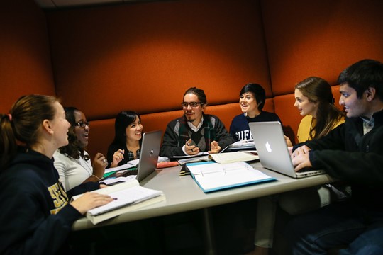 International students at INTO Suffolk University sit at large desk with laptops, notebooks, and books open, collaborating on group project. 