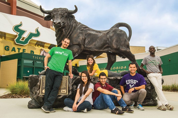 Six international students from INTO University of South Florida pose in colorful university and INTO clothing in front of statue of Rocky the Bull at the USF Yeungling Center.