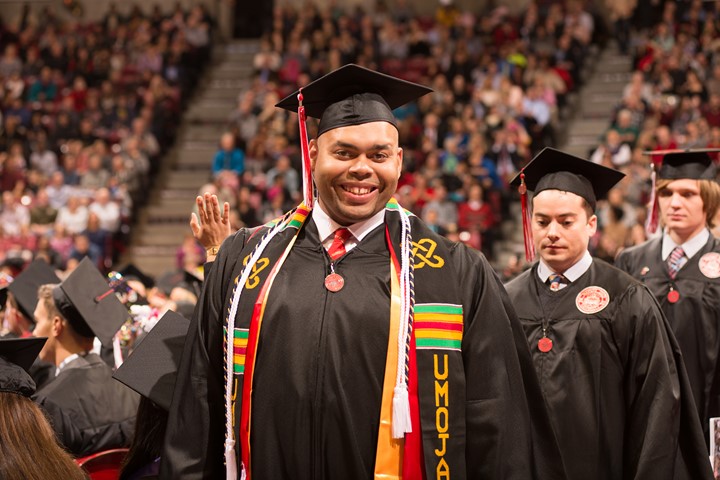 INTO Illinois State University international student smiles to camera while wearing cap, gown, and honors stoles at graduation ceremony in arena.