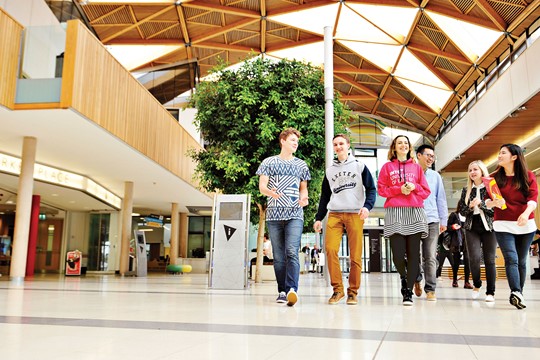 INTO University of Exeter international students laugh as they walk through forum with wood paneled, triangular patterns on ceiling.