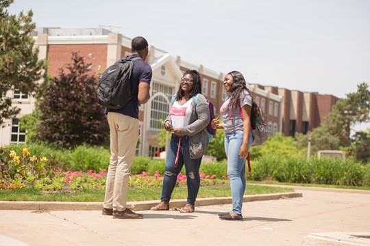 Three international students from INTO Illinois State University stop and talk on campus pathway near bright pink flowers and brick building.