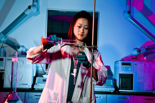 International student at INTO Drew University conducts experiment involving beaker while wearing white coat and latex gloves in blue-lit laboratory. 