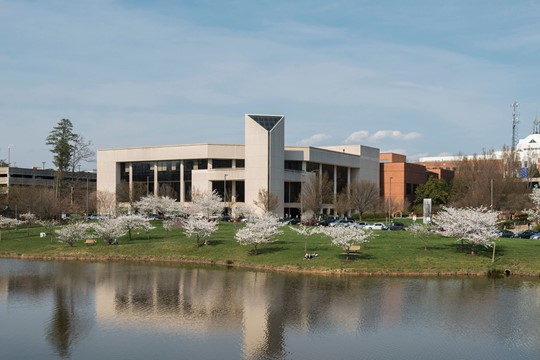 Exterior view of George Mason University building, with lawn, trees with white blossoms, and pond in foreground, and other buildings in background.