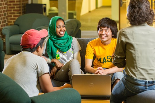 Group of four international students gather and speak on couches and chairs around laptop in communal area of INTO George Mason University Center. 