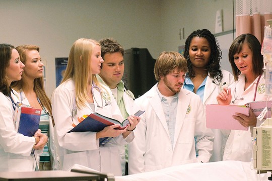 Group of medical students at The University of Alabama at Birmingham wear white coats and look at chart held by instructor near patient bed.