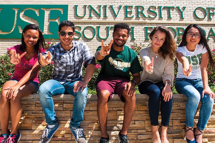 Five international students from INTO University of South Florida sit and smile in front of USF sign and flower bushes while making bull horns with right hands.