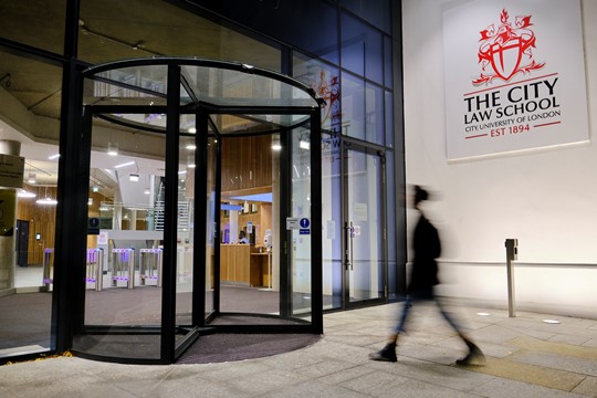 Student, blurred in motion, walks toward rotating doors of City, University of London Law School, the exterior of which is illuminated at night.