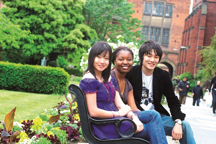 Three INTO Newcastle University international students sit and smile on bench adjacent to green lawns and in front of historic, brick buildings.
