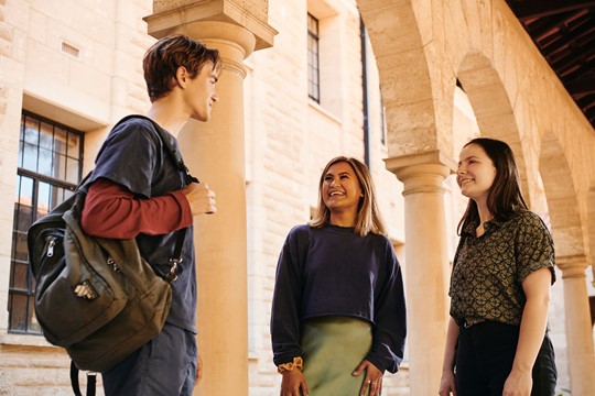 Three students of The University of Western Australia stand beneath arches and smile while speaking to each other on campus.