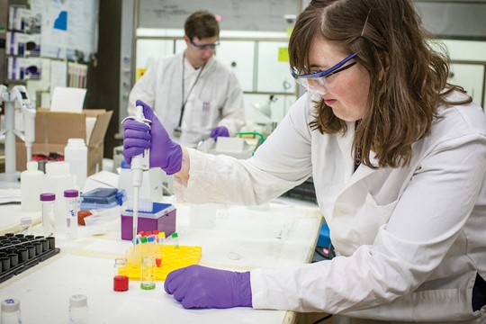 Oregon State University students conduct experiment involving multiple glass vials in laboratory while wearing white coat, latex gloves and goggles.