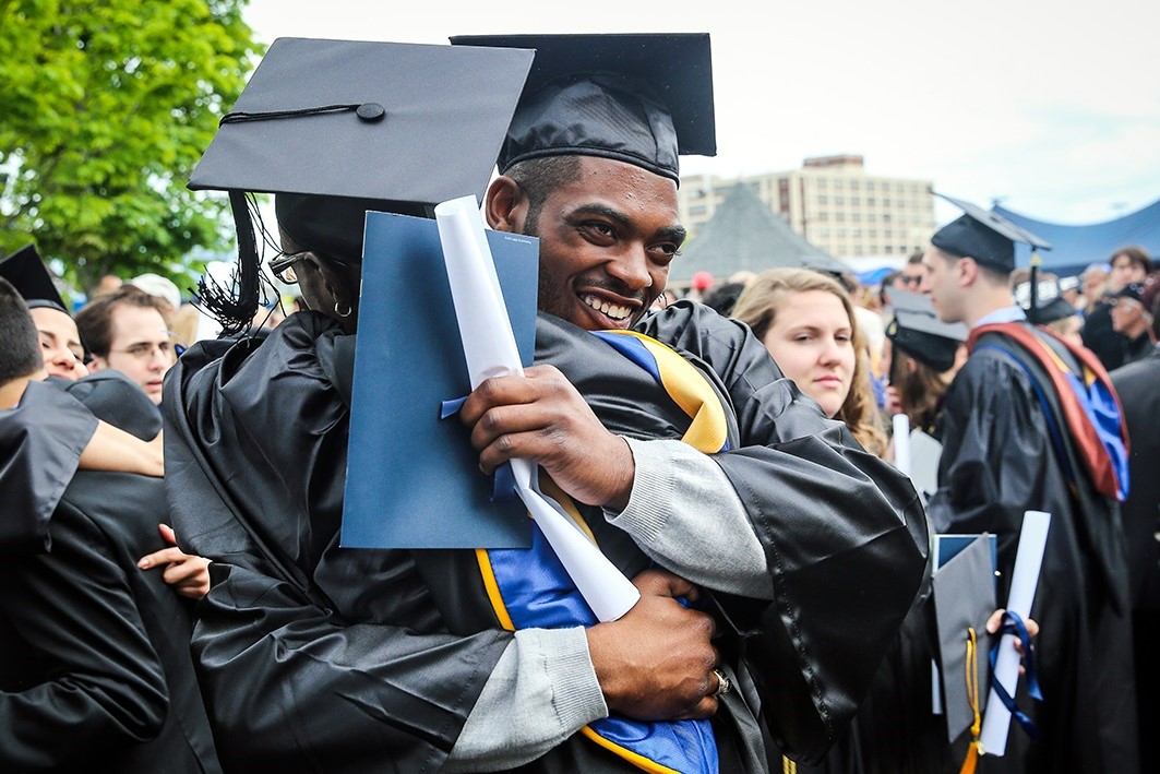 International student from INTO Suffolk University in cap and gown embraces peer at graduation ceremony.