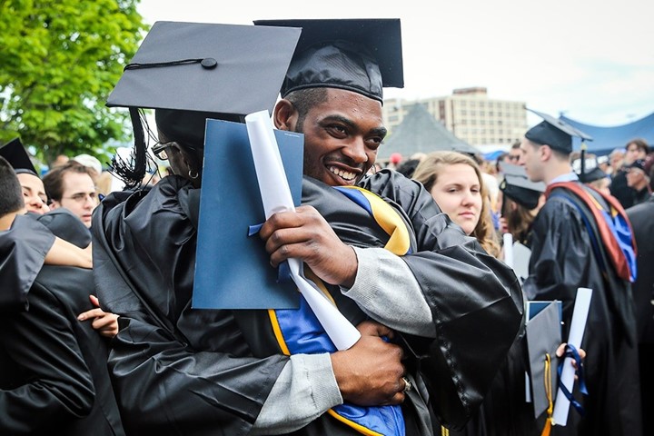 International student in cap and gown smiles and embraces friend while holding a diploma at Suffolk University commencement ceremony.