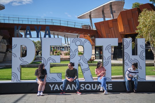 Four students of The University of Western Australia sit interspaced in front of artistic statue spelling out “Kaya Perth,” in Yagan Square.