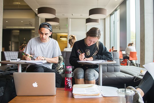 Two students sit on couch and write on worksheets on mobile desks at INTO Oregon State Living and Learning Center. Laptop and notebook on table in foreground.