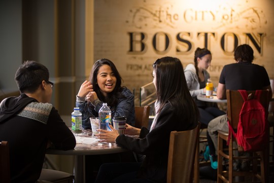 Three international students at INTO Suffolk University sit and talk at wooden table in cafeteria in front of wall that reads “The City of Boston.”