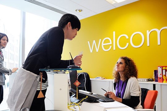 International student speaks to receptionist at INTO City, University of London Centre information desk in front of yellow wall and “welcome” sign.