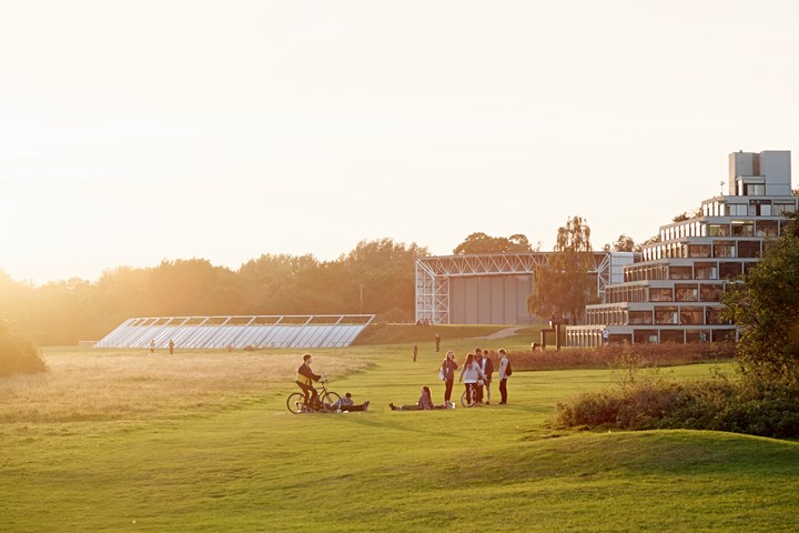 Students gather on green lawns of University of East Anglia’s Norwich campus at dusk, with UEA’s landmark ziggurats in background.