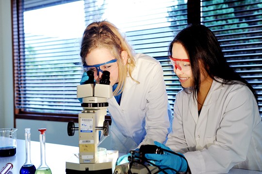 Two INTO University of Stirling international students look into microscope while wearing goggles, white coats, and latex gloves in laboratory.