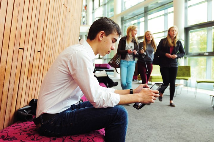 International student sits on pink, floral-patterned bench and works on a tablet in hallway of INTO University of Exeter building as three students walk by.