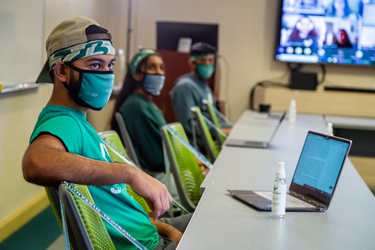 Three international students practice social distancing and wear masks in INTO University of South Florida Center classroom with laptops and hand sanitizer.