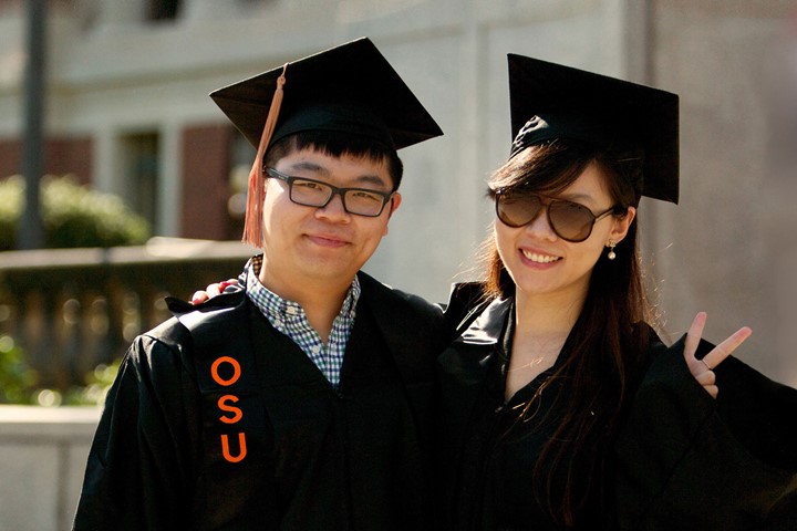 Two international students from INTO Oregon State University wearing caps and gowns, smile as they celebrate graduating from university. One makes peace sign with hand.