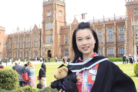 INTO Queen’s University Belfast international student smiles and poses in cap and gown holding teddy bear in front of Lanyon Building on Queen’s campus. 