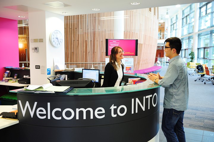 International student speaks to receptionist at INTO University of Exeter building information desk. Words “Welcome to INTO” visible on front of desk.