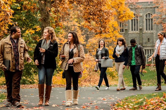 Students walk down pathway on Drew University campus on fall day, with trees with yellow and orange leaves and stone building in background.