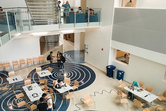 Multi-level science building interior, with students at desks designed as periodic table squares on first floor and more attending lecture on second floor. 