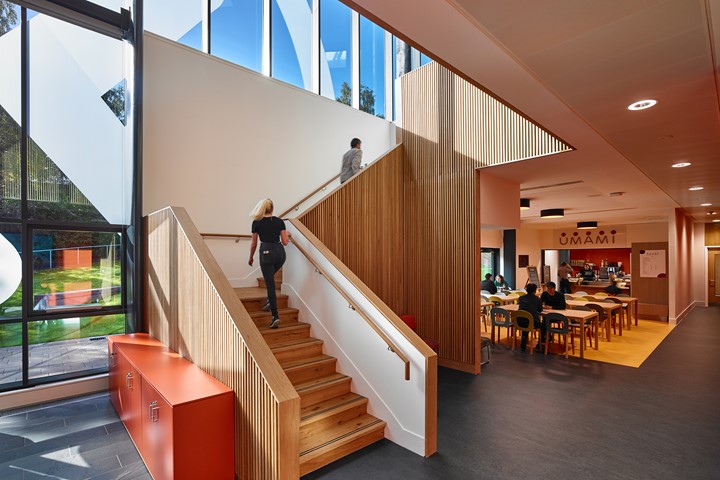 Two international students climb a wooden staircase outside of busy Umami Café in bright INTO campus, with windows in background.