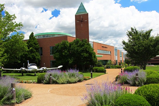 Parks College of Engineering, Aviation and Technology Building with brick clock tower, two-seater airplane, and walkway lined with bushes in foreground. 