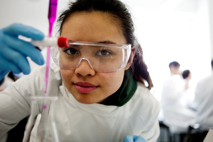 Close-up view of international student conducting experiment with test tube while wearing white coat, latex gloves and protective glasses.
