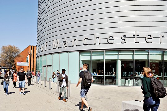The University of Manchester students walk on pavement in front of University Place, also known as the Tin Can, a round, metallic building. 