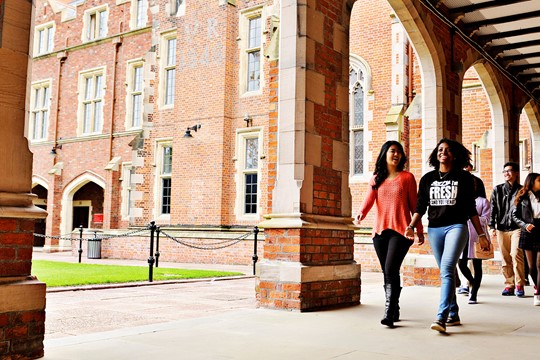 Queen’s University Belfast students walk on pavement in front of brick arches, brick buildings, and lawn on campus.